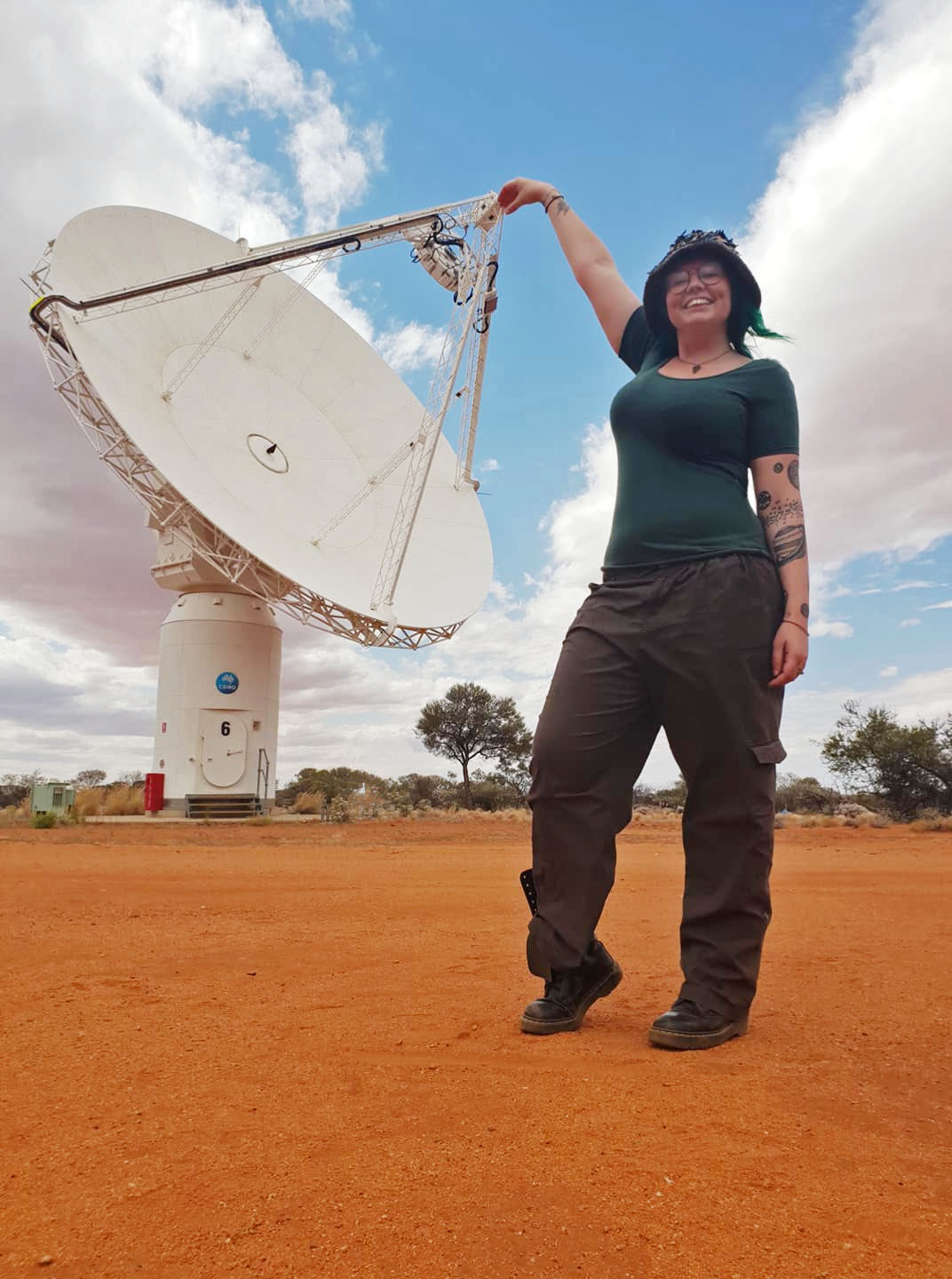 A woman stands infront of a large radio telescope in a desert landscape